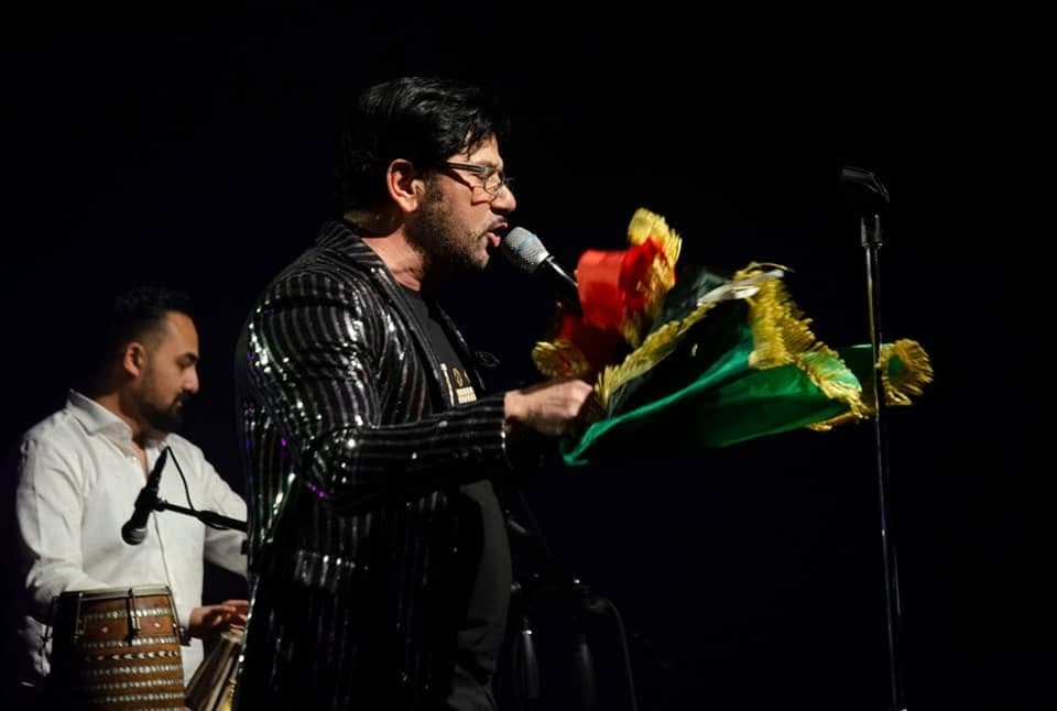 Taher Shubab in a sequinned jacket singing on a dark concert stage
